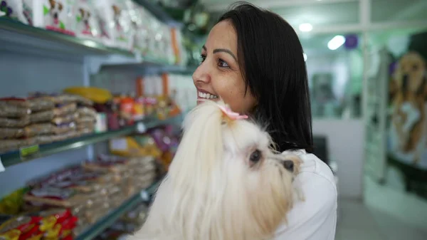 Woman standing inside Pet Shop business observing products on shelf while holding small dog in arms