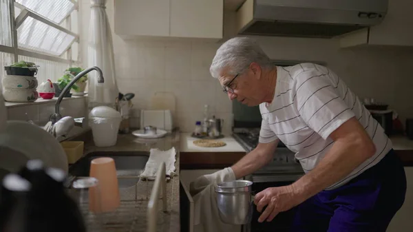 キッチンに立つシニア男性は自宅で窓ごと鍋やフライパンを整理シンクします 老人の本物の家庭シーン — ストック写真