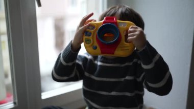 Elinde oyuncak fotoğraf objesiyle fotoğraf çekiyormuş gibi yapan çocuk. Eğlenceli küçük çocuk evde yaratıcı eğlencelerle oynuyor.