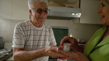 Yaşlı bir adam termosla kahve servisi yapıyor mutfakta duruyor ve karısına sıcak bir içecek veriyor.