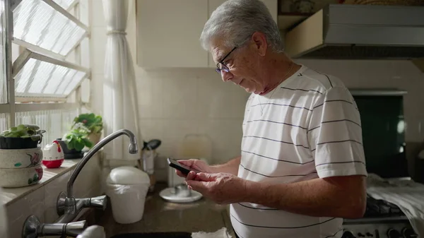 Hombre Mayor Mirando Dispositivo Teléfono Móvil Mientras Está Pie Cocina —  Fotos de Stock