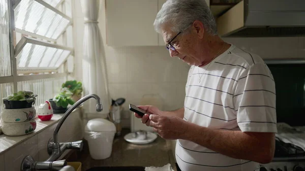 Äldre Man Tittar Mobilen När Han Står Köket Vid Fönstret — Stockfoto