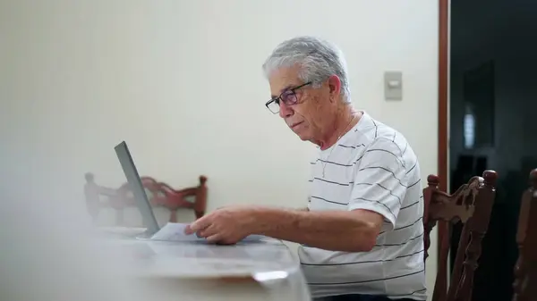 一位心烦意乱的老人坐在电脑前 不得不花钱买汽车 坐在家里厨房里 用笔记本电脑和键盘打字 — 图库照片