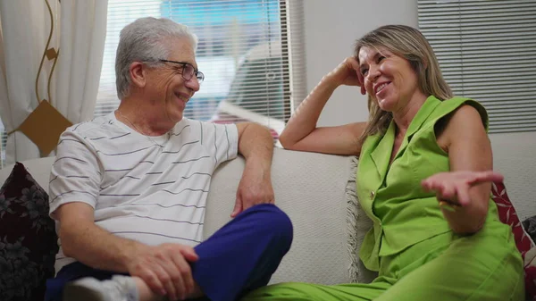 Happy mature man and woman conversing seated on couch at home indoors. Senior man and middle-aged female caucasian woman interacting, smiling and laughing together