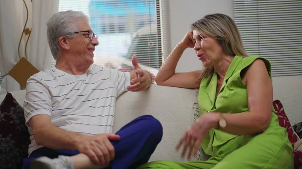 Happy mature man and woman conversing seated on couch at home indoors. Senior man and middle-aged female caucasian woman interacting, smiling and laughing together