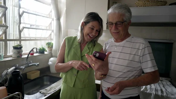 Wife sharing phone screen to husband while standing in kitchen. Middle-age couple laughing and smiling while watching content online with modern technology