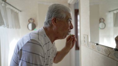 Sabah rutininin bir parçası olarak dişlerini fırçalayan yaşlı adamın sıradan bir sahnesi. Yaşlı insan diş hijyeni ve yüz yıkama, güne başlama ritüeli