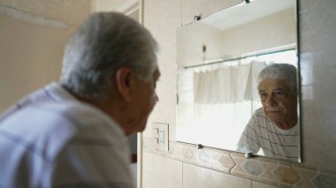 Yaşlı beyaz erkek, banyo aynasında kendi yansımasına bakıyor. Yaşlı, kır saçlı, yaşlılıktan pişmanlık ve üzüntü duyan yaşlı bir adam.