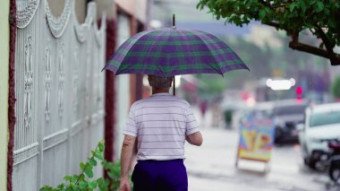Elinde şemsiyeyle yağmurda yürüyen son sınıf öğrencisinin arkası. Yaşlı yetişkin insan yağmur yağarken kaldırımda geziniyor.