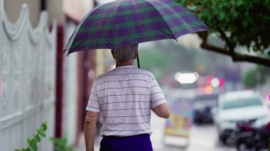 Elinde şemsiyeyle yağmurda yürüyen son sınıf öğrencisinin arkası. Yaşlı yetişkin insan yağmur yağarken kaldırımda geziniyor.