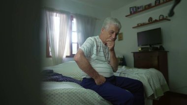 Düşüncelere dalmış yatağın yanında oturan düşünceli kıdemli bir adam. Emekli yaşlı insan, kararlarını el pençe divan düşünerek gündelik yatak odasında derin bir zihinsel yansıma içinde düşünür.