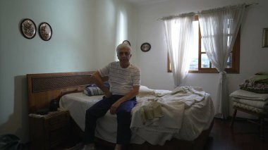 Yatağının yanında oturan yaşlı bir adam, rahat bir yatak odasında beyaz saçlı yaşlı bir adamın otantik gerçek hayat portresi, yaşlılık emekliliğini resmeden bir yaşam tarzı.