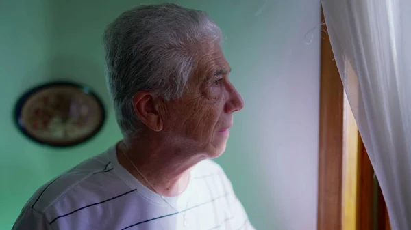 自宅の窓から立って 年賀状のカーテンを通して眺めているシニアマン 居住地から近所を眺めている高齢者グレー ヘアマン — ストック写真