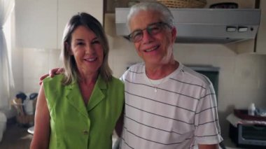 Mutlu yaşlı çift kameraya poz veriyor. Mutfakta duruyor, orta yaşlı evli erkek ve kadın gülümsüyor.