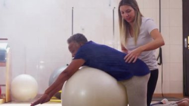Kadın spor koçu, fizyoterapi seansı sırasında siyahi bir kadına pilates topuyla egzersiz yapması için yardım ediyor.