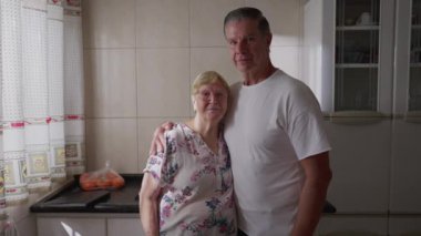 Yaşlı anne ve yetişkin orta yaşlı oğul kameraya birlikte poz veriyorlar, mutfak penceresinin önünde duruyorlar.
