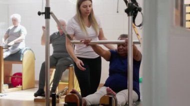 Kadın koç, siyah yaşlı bir kadına pilates makinesini kullanması için yardım ediyor. Spor eğitmeni, yaşlılık egzersizleri öğretiyor. Sağlıklı yaşlı yaşam tarzı.
