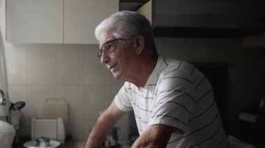 Kederli yaşlı adam pencerenin kenarında duruyor ve pişmanlık ve endişe duyuyor. Evde yalnızlık çeken, kafası karışık, gri saçlı bir erkek.