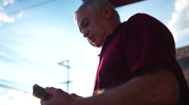 Üst düzey bir adam dışarıda parlayan güneş ışığıyla duran cep telefonu cihazına bakıyor. Akıllı telefon ekranına bakarak modern teknolojiyle ilgilenen yaşlı kişi