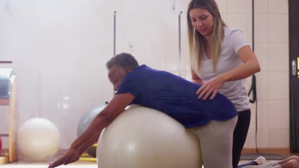 女性スポーツコーチは 理学療法セッション中にピラティス ボールと運動する黒人シニアの女性を支援 — ストック動画
