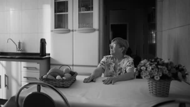 孤独のシニア ウーマン モノクロのキッチン テーブルに座った 劇的な本物の高齢者のおばあちゃんは思考で失われた見つめている — ストック動画