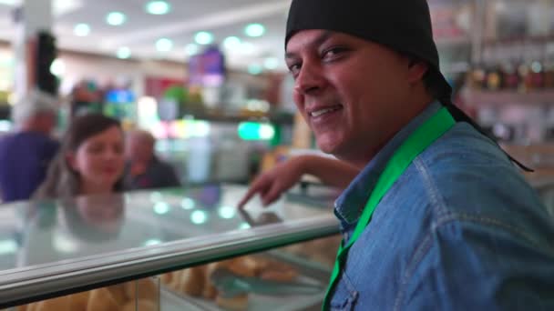 カウンターの後ろに立っている食料品店の喜びの従業員は食糧順序を選ぶ顧客と対話しながら笑顔のカメラに頭を回します — ストック動画