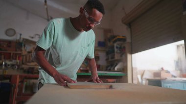 Genç bir siyah marangoz atölyede odun kesmek için makine kullanıyor. Marangozluk işine odaklanmış titizlikle çalışıyor.