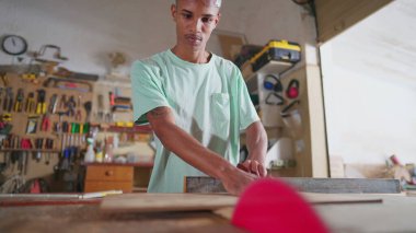 Genç bir siyah marangoz öğrenci ahşap ölçer, testere makinesini mezura ile kullanmaya hazırlanıyor. Atölyede konsantre marangoz, öğrenim mesleği