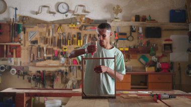 Bir genç siyah marangoz marangozluk işyerinde çalışıyor, ahşap yüzey mobilyalarına yapıştırıcı sürüyor ve ahşap parçaları yapıştırıyor.
