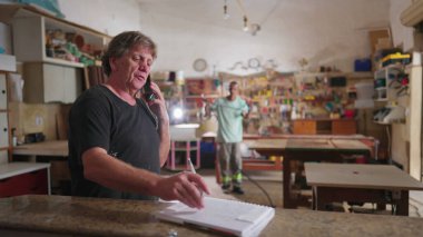 Marangozhanenin mutlu işletme sahibi müşteriyle telefonla konuşuyor ve araçlarla arka planda çalışan çırağına onay veriyor.