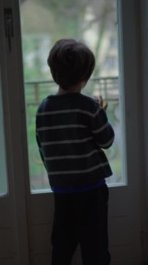 Melankolik küçük çocuk pencereden manzaraya bakıyor. Düşünceli çocuk birinin gelmesini bekliyor, dikey video.