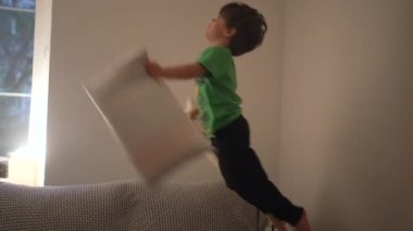 Çocuk akşamları kanepede zıplayıp eğleniyor. Küçük çocuk yastıklarla kanepeye dalıyor.