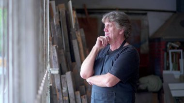 Düşünceli, yaşlı bir marangoz sokak kenarındaki atölyede durup derin düşüncelere dalmış bir ifadeyle dışarı bakıyor. Gri saçlı küçük işletme sahibi kararını gözden geçiriyor.