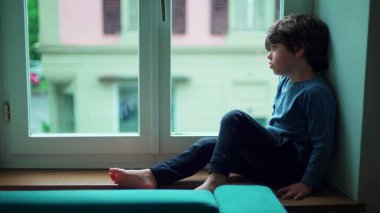 Canı sıkılan küçük bir çocuk, pencerenin kenarında oturuyor ve can sıkıntısı çekiyor. Çocuk eve kapanmış, huzursuz hissediyor.