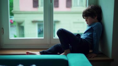 Canı sıkılan küçük bir çocuk, pencerenin kenarında oturuyor ve can sıkıntısı çekiyor. Çocuk eve kapanmış, huzursuz hissediyor.