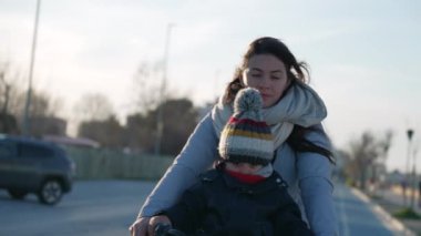 Anne ve 2 yaşındaki çocuk soğuk kış gününde bisikletle dışarıda. Eşarp ve ceket giyen çocuk ve ebeveyn bisiklet sürüyor.