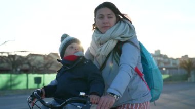 Anne, soğuk kış gününde arka planda güneş ışığı parlarken çocukla birlikte bisiklete biner. Annem bisiklete biniyor