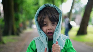 Kapüşonlu ceketli dört yaşındaki çocuk yeşil yolda dikiliyor. Moda giysiler giyen küçük bir çocuğun yakın plan yüzü.