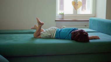 Sıkılmış çocuk yapacak bir şey olmadan kanepede yatıyor, huzursuz küçük çocuk evde oturmuş sıkıntıyla boğuşuyor.