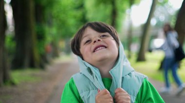 Kapüşonlu ceketli dört yaşındaki çocuk yeşil yolda dikiliyor. Moda giysiler giyen küçük bir çocuğun yakın plan yüzü.