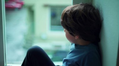 Pencere kenarında oturan düşünceli küçük çocuk dışarı bakıyor, küçük çocuğun düşünceli düşünceli bakışları.