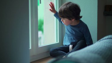 Pencerenin kenarında oturan bir çocuk sokağa bakıyor, cama yaslanmış evden manzaraya bakıyor.