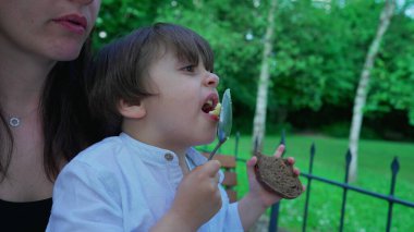 Çocuk, açık hava restoranında annesinin kucağında kaşıkla püre yemeği tadıyor. Uzaktan bakan, yemek yiyen çocuğun yakın plan yüzü.