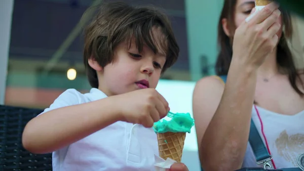 Child eating colorful ice-cream cone with mom outside. One caucasian little boy enjoys sweet dessert during summer day
