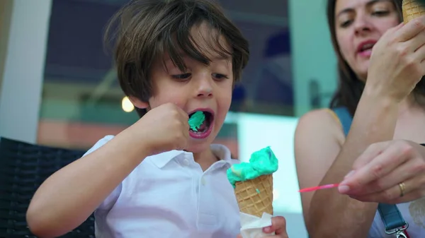 Child eating colorful ice-cream cone with mom outside. One caucasian little boy enjoys sweet dessert during summer day