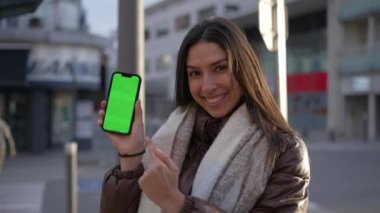 Neşeli bir Orta Doğulu genç kadın şehir caddesinde dururken elinde krom renkli yeşil ekran bir cep telefonu ekranı tutuyor. Kadın kişi çevrimiçi video içeriğini işaret ediyor