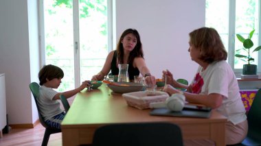 Evde samimi bir aile hayatı, yetişkin kadın ve çocukla sıradan bir yaşlı kadın yemek masasında oturuyor.