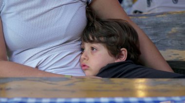 Bored little boy clinging to mother's body with nothing to do, child struggles with boredom while holding into parent gazing at distance daydreaming clipart