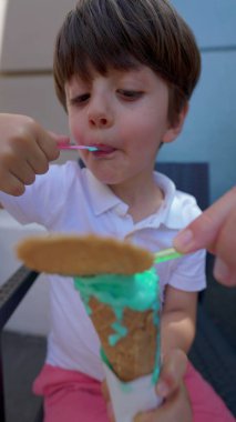 Küçük çocuk güneşli yaz gününde renkli dondurma külahıyla kendini şımartıyor. Tatlı yiyen çocuk, dikey video.