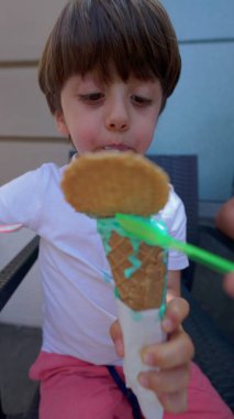 Küçük çocuk güneşli yaz gününde renkli dondurma külahıyla kendini şımartıyor. Tatlı yiyen çocuk, dikey video.